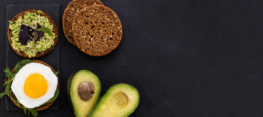 Avocado sandwich on dark rye bread with spinach and fried egg on a dark background, top view with copy space.