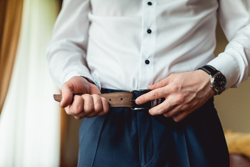 Groom in blue pants and a white shirt fastens his leather belt. grooms mourning. Wedding details.