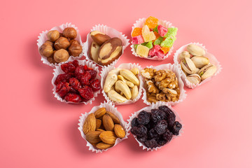 A serving of assorted candied fruit, dried cherries, almonds, raisins, walnuts and hazelnuts in paper muffin cups on a pink background.