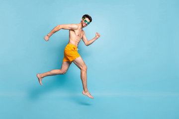 Full length body size profile side view of nice attractive cheerful funky guy in swimming shorts jumping running having fun isolated on bright vivid shine vibrant green blue turquoise color background