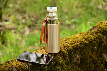 Photo of a thermos and smartphone in the forest in the summer outdoors. Tourist vacation concept.