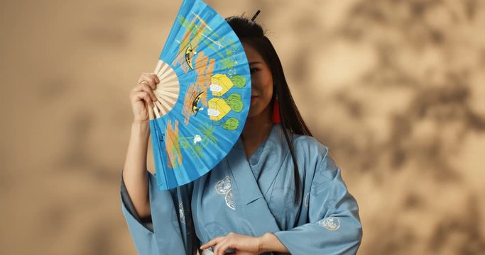 Portrait shot of beautiful young geisha in blue kimono waving with fan and closing her face while smiling and flirting. Pretty Asian woman in traditional outfit.
