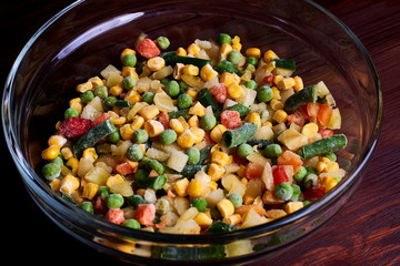 Frozen different vegetables in a glass plate on a wooden background.