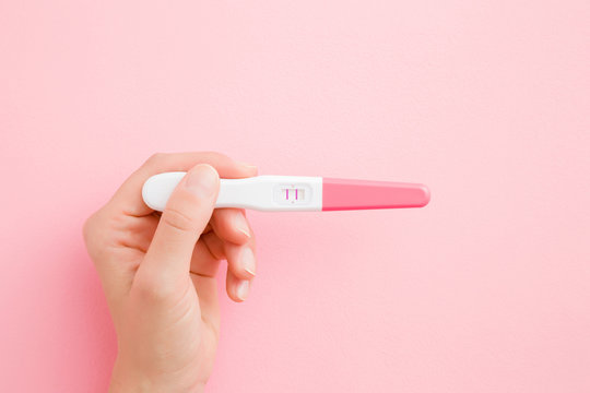 Young woman hand holding pregnancy test with two stripes on light pink table background. Pastel color. Positive result. Closeup. Point of view shot. Top down view.