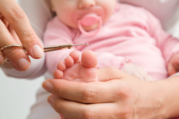 Young mother hand holding infant foot and cutting toenail with baby scissors. Closeup. Front view.