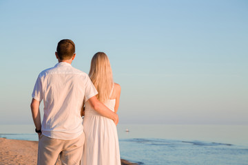 Young couple in white clothes. Man hugging woman at beach in summer evening. Orange sunset light. Back view. Empty place for inspirational, sentimental text, positive, lovely quote or cute sayings.