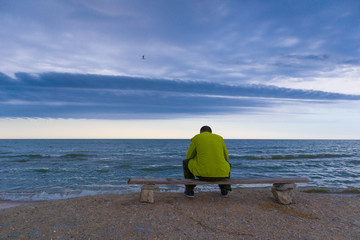 Man sitting on a bench near a beach and looking on the ocean with epic clouds. 