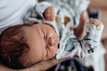 Candid real family moment. Soft focus. Young dark hair father holding his newborn baby son sitting in a bedroom. Boy is wearing body with watercolor animal print. Father's day. Boy is sleeping.