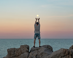 Young man in casual clothes stands on the rocks by the sea at sunset and the full moon between his arms raised 