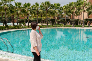 A middle-aged woman, during a coronavirus, in a mask, talking on the phone, near the pool, in the hotel.