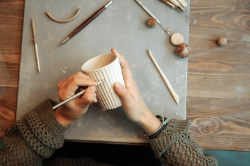 Top view of hands with clay cup and sculpting tool. Working in a pottery workshop. Hobby.