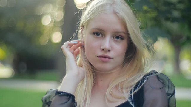 Attractive happy schoolgirl in a black translucent blouse walks in nature, confidently poses and straightens her long chic blond hair. A young female model is shyly smiling and looking up.