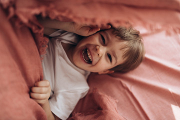 Soft focus. Look like film shot. Four years boy is lying on the back on terracotta bed linen bed in white shirt and smiling