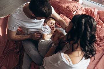 Pregnant mother playing with her child in the bedroom. Mother, father and their son are sharing good emotions while relaxing at home Big window, natural light, terracotta bed linen. Family at home.