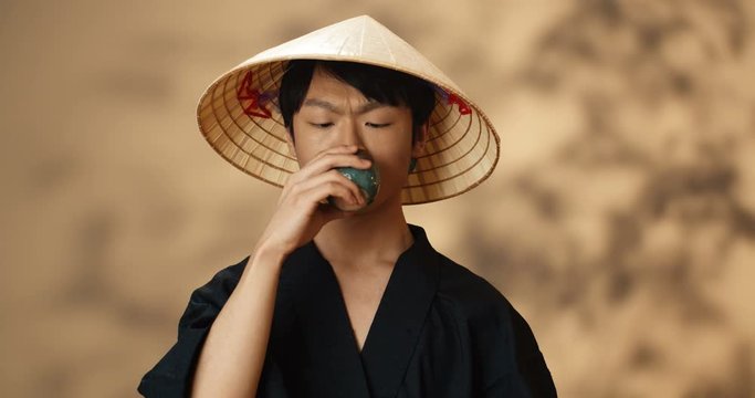Portrait of happy handsome Asian young man in conus hat sipping hot drink from ceramic cup and smiling cheerfully to camera. Guy drinking tea. Chinese tea ceremony concept.