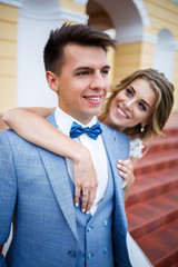 Young stylish guy in the costume of the groom and the bride beautiful girl in a white dress with a train walking on the background of a large house with columns on their wedding day