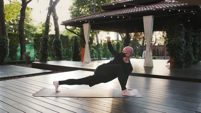 Strong barefoot guy in black protective mask and sport clothes is practicing yoga on mat in courtyard. Healthy lifestyle during Coronavirus pandemic