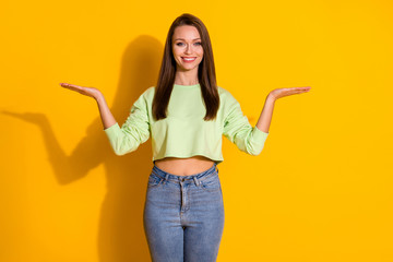Photo of attractive pretty lady good mood hold open arms empty space two novelty products choose best one wear green cropped sweatshirt jeans isolated vivid bright yellow color background