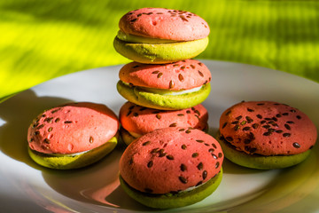 Multi-colored green and pink, bright cookies with two halves dressed with filling and sprinkled with sesame seeds on a white plate and on a bright salad background.