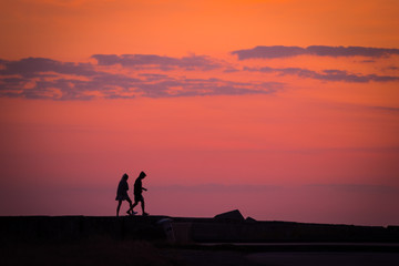 Fototapeta na wymiar Pareja caminando en el ocaso de un hermoso dia de verano durante junio, julio o agosto junto al mar bajo el color de las nubes y sus tonos naranjas, rojos o morados 