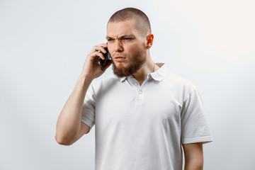 Portrait of an upset handsome bearded man in a white t-shirt talking on a smartphone. isolated, emotions