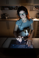 A smiling woman in headphones sits in the dark at a wireless computer in the kitchen with a puppy of Jack Russell Terrier on her knees. Girl with her little dog watching a movie on a laptop at home.