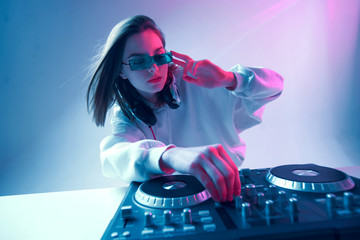 Cool young girl DJ mixes music on a mixing console and headphones, in stylish clothes, glasses on a...