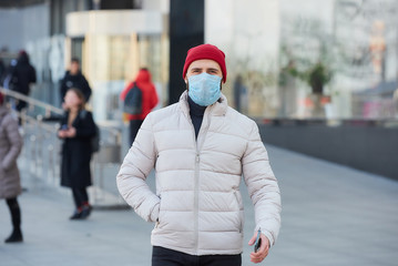 A man with a beard wearing a medical face mask to avoid the spread coronavirus (COVID-19). A guy with a surgical mask on the face because of the pandemic walking with a smartphone down the street.
