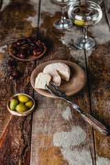 Cheese, olives and dates in plates with glass of martini drink on brown wooden table, selective focus