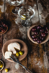 Cheese, olives and dates in plates with glass of martini drink on brown wooden table, selective focus