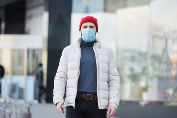 A man in a red watch cap wearing a medical face mask to avoid the spread coronavirus (COVID-19). A guy with a surgical mask on the face because of the pandemic walking in the center of the city.