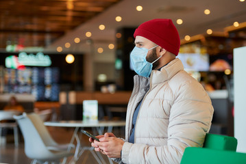 A caucasian man with a beard wearing a medical face mask to avoid the spread coronavirus (COVID-19). A fellow with a surgical mask on the face holding a smartphone in the shopping center.