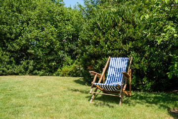 A deck chair in a nice garden surrounded by green bushes on a beautiful sunny day.