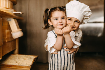 Little boy hugging tight little girl, smiling. Brother and sister wearing cook clothes in kitchen.