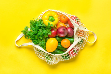 Full eco mesh bag of different health food - bell pepper, tomatoes, bananas, lemon, green, mandarin, cucumber, onions on yellow background Top view