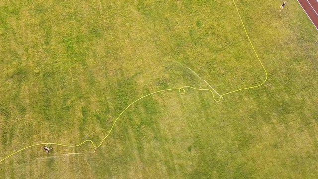 Top down aerial view of football field surface covered with green grass and sprinklers spraying water.
