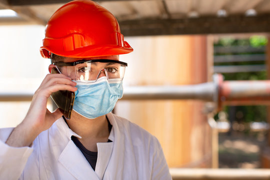 Tecnico specialista in camice bianco e caschetto protettivo arancione indossa una mascherina chirurgica e degli occhiali tecnici , parla al telefono  in contesto industriale.