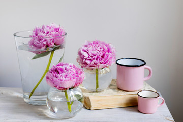 Three pink peonies in round vases on a beige wooden table against the background of a white wall.
