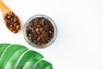 Body scrub from ground coffee and sugar in a glass jar, coffee beans in a wooden spoon and a leaf of monstera on a white background. Homemade peeling and spa care product. Top view.