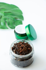 Body scrub from ground coffee and sugar in a glass jar, a green leaf of monstera and face cream on a white background. Homemade peeling and spa care product.