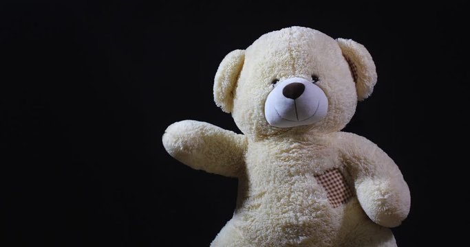 A lively toy bear looking at the camera and waving its paw as a sign of welcome on a black studio background with dramatic light and free space for inscription.