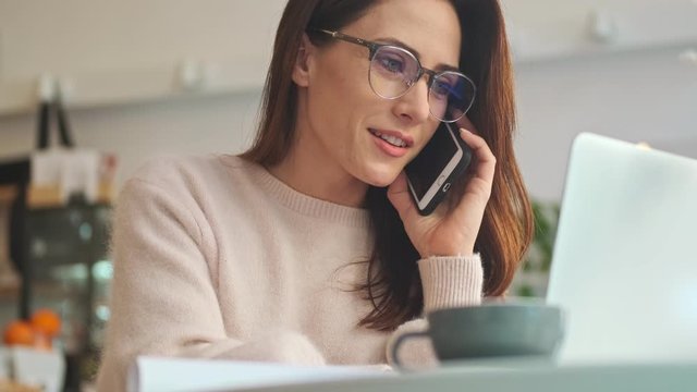 A smiling young woman wearing glasses is working while talking on the phone in the cafe indoors