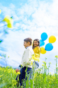 Excited kids with balloons run in field.Jump and fly in blooming yellow rape. blue sky, green grass. Summer beautiful card. Freedom concept.
