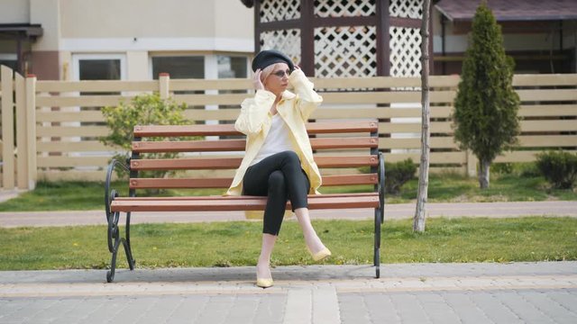 Wide shot of elegant blond woman putting on sunglasses, standing up from bench and leaving. Portrait of confident Caucasian lady resting outdoors on sunny day. Elegance, confidence, beauty, lifestyle.
