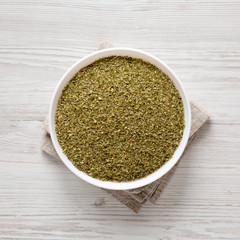 Dried Green Greek Oregano Spice in a white bowl on a white wooden surface, top view. Flat lay, overhead, from above.