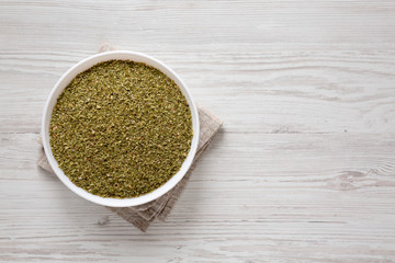 Dried Green Greek Oregano Spice in a white bowl on a white wooden surface, top view. Flat lay, overhead, from above. Copy space.