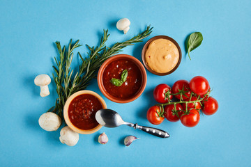 top view of delicious sauces in bowls with spoon near fresh ripe vegetables on blue background