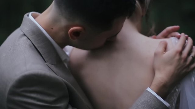 Close-up of a young groom in a suit stroking and kissing his bride in the back in a wedding dress outside in nature. Romantic getaway of a happy cute european honeymoon couple in spring honeymoon.
