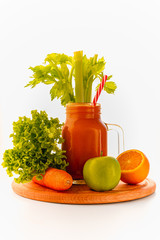 Glass mason jar of orange and carrot smoothie with  a drinking straw standing on a round wooden kitchen shelf surrounded by leaves, carrot, a green apple and an orange

