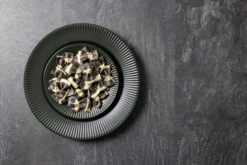 Italian farfalle black pasta, on a dark background stone, black plate. Top view. Copy space.